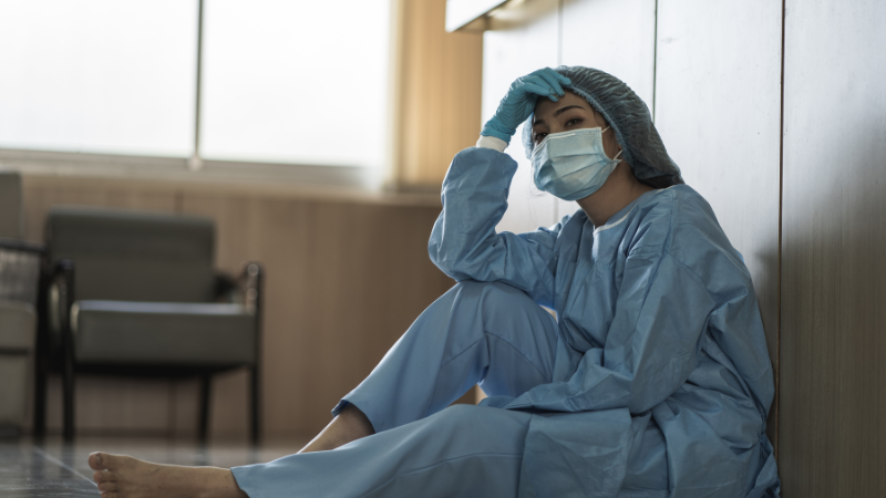 A Nurse Practitioner wearing scrubs, mask, and hair cover sits on the floor looking fatigued, with hand on the head, grappling with burnout in a hospital setting.