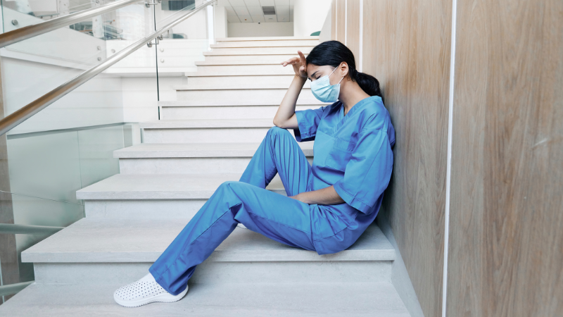 A nurse practitioner in blue medical scrubs and a face mask sits on stairs, appearing fatigued and resting their head in their hand, trying to handle the ongoing burnout.