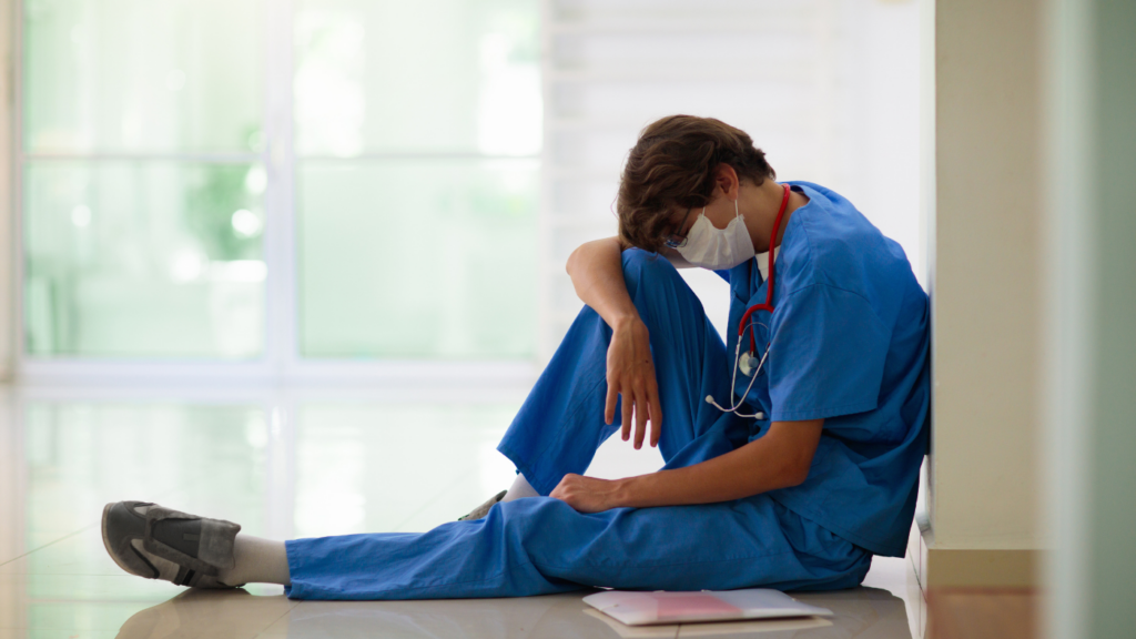 A tired healthcare worker in blue scrubs and a face mask sits on a hospital floor, head down, with a stethoscope around their neck and a notebook on the ground nearby, embodying the risk factors associated with nurse practitioner burnout.