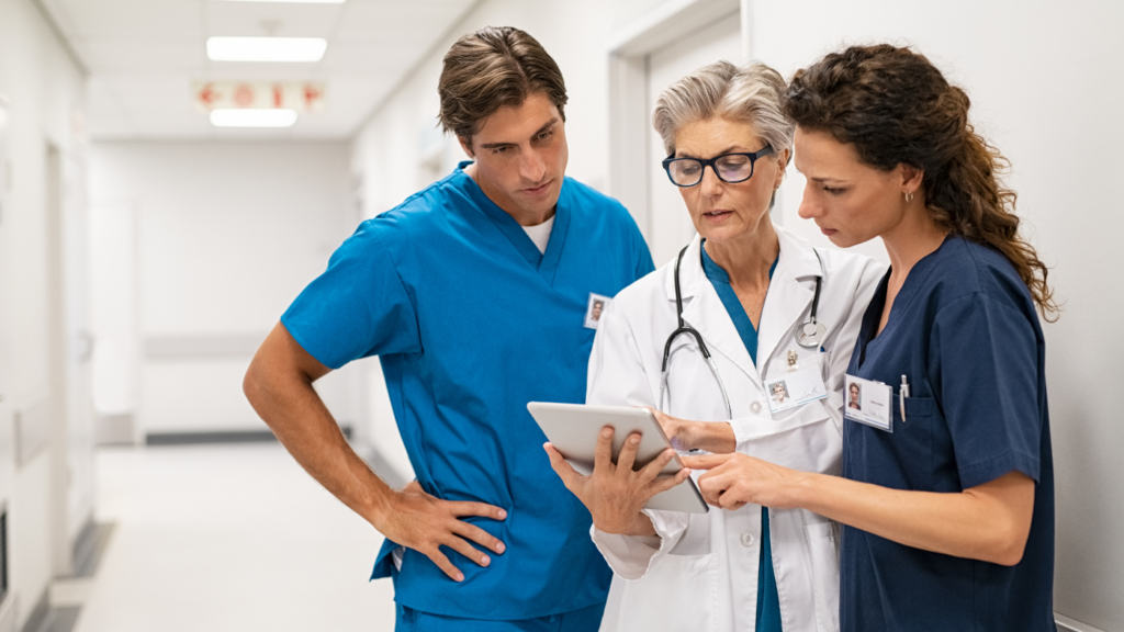 Three healthcare professionals in scrubs and lab coats discuss patient information using a tablet in a hospital hallway, highlighting their Nurse Practitioner specialty.