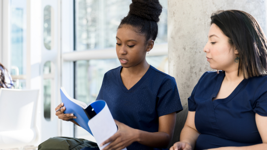 Two women in blue scrub uniforms are sitting together; one is holding and reading from a folder while the other listens attentively. They are indoors, near a window, likely discussing strategies to ace their NP Certification Exam.