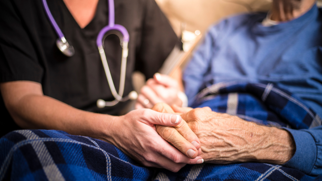 A Nurse Practitioner specializing in Adult-Gerontology Primary Care holds hands with an elderly patient, who is comforted by a blue plaid blanket.