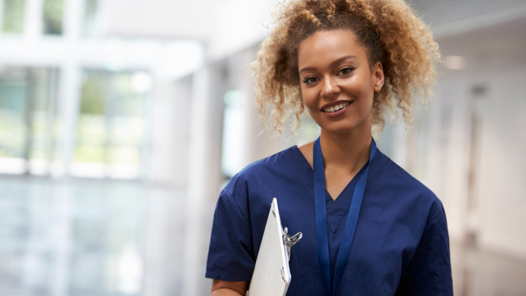 A Nurse Practitioner in blue medical scrubs and a lanyard holds a clipboard and smiles while standing in a bright, modern hallway, confidently battling Impostor Syndrome.
