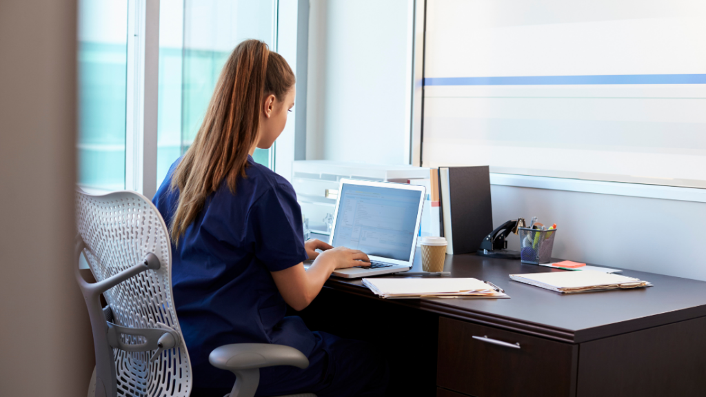 A person in medical scrubs sits at a desk working on a laptop, surrounded by office supplies, a coffee cup, and files in a modern office. Despite the professional environment, they battle imposter syndrome as they meticulously go through their tasks.