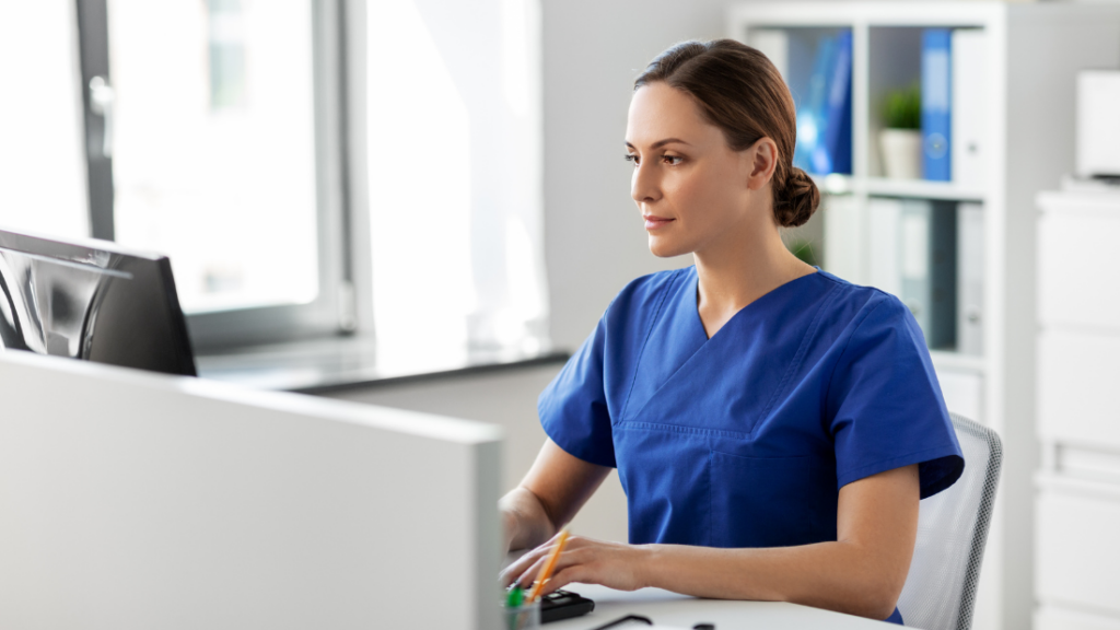 A person wearing blue medical scrubs is working at a desk on a computer in a well-lit office, showcasing the diverse scope of practice nurse practitioners handle daily.