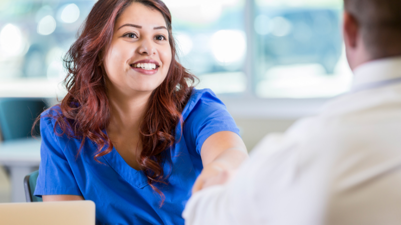 A woman in blue scrubs, likely a nurse practitioner, is smiling and shaking hands with a man during an informal job interview in a brightly lit room.