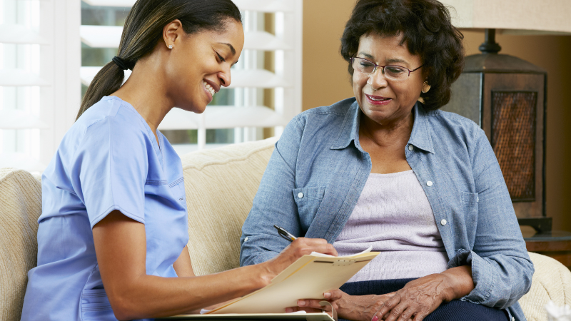 A Women's Health Nurse Practitioner in blue scrubs sits and writes on a clipboard while conversing with an elderly woman wearing a light gray shirt and denim jacket on a sofa.