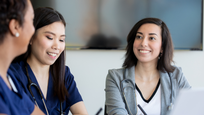 Three healthcare professionals, two wearing scrubs, are seated and conversing while smiling in a meeting room, possibly discussing NP Board Prep strategies. Stethoscopes are visible around their necks.
