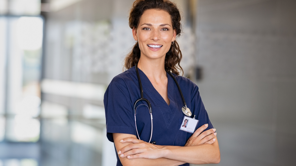 A healthcare professional with a stethoscope around her neck and a name badge, wearing blue scrubs, stands in a modern medical facility, smiling at the camera with arms crossed. Her demeanor reflects the expertise gained through an advanced practice nursing degree.