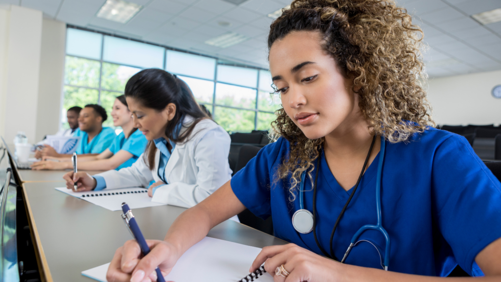 A group of medical students in scrubs and lab coats take notes in a classroom, gearing up for their nursing career. A woman in blue scrubs with curly hair is in the foreground, writing diligently in a notebook, perhaps reflecting on her experiences from the nurse practitioner program.