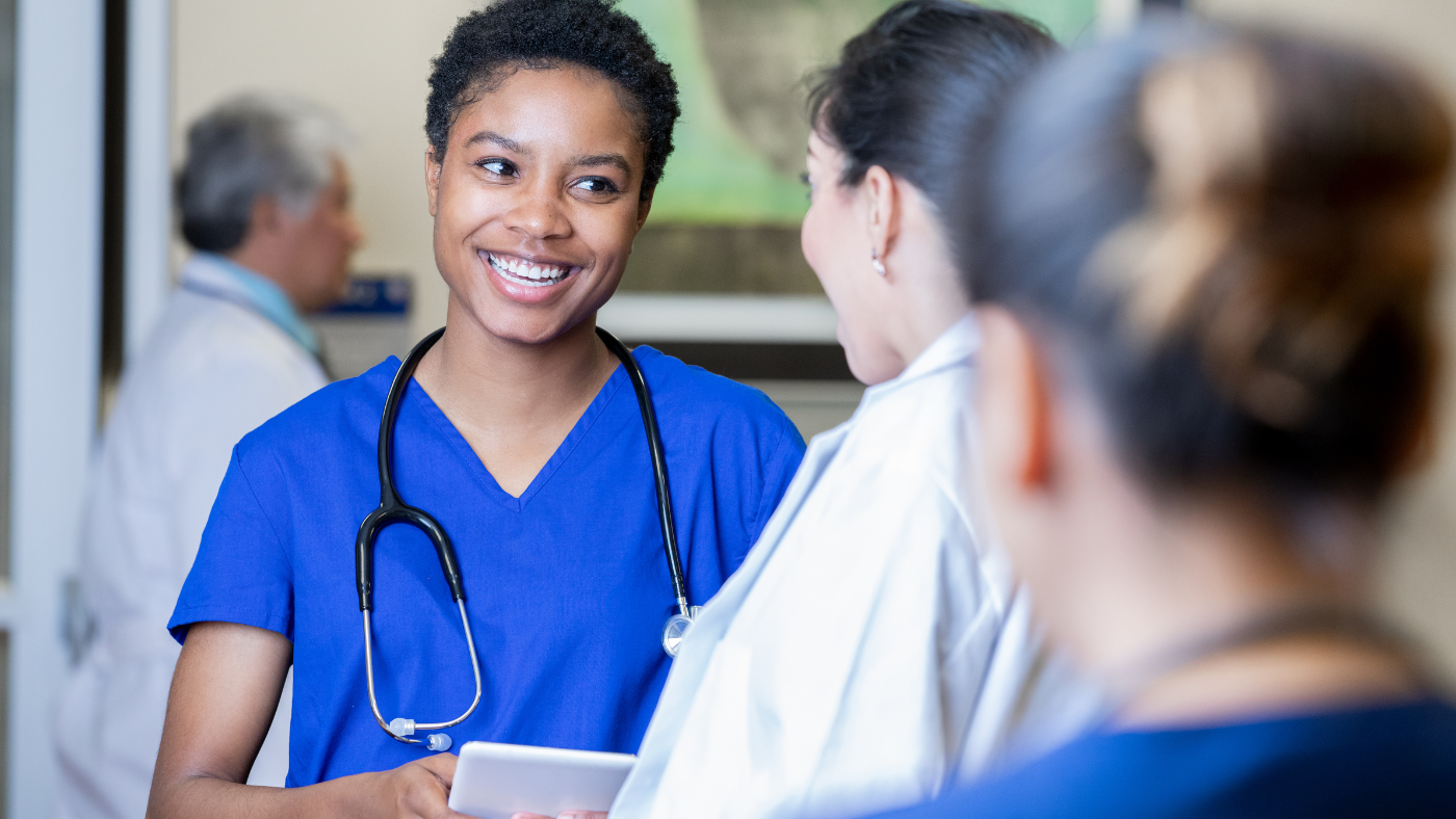 A medical professional in blue scrubs smiles while talking to a colleague, finding balance in their workday. Another person in the foreground has their back to the camera, while a different individual is blurred in the background.