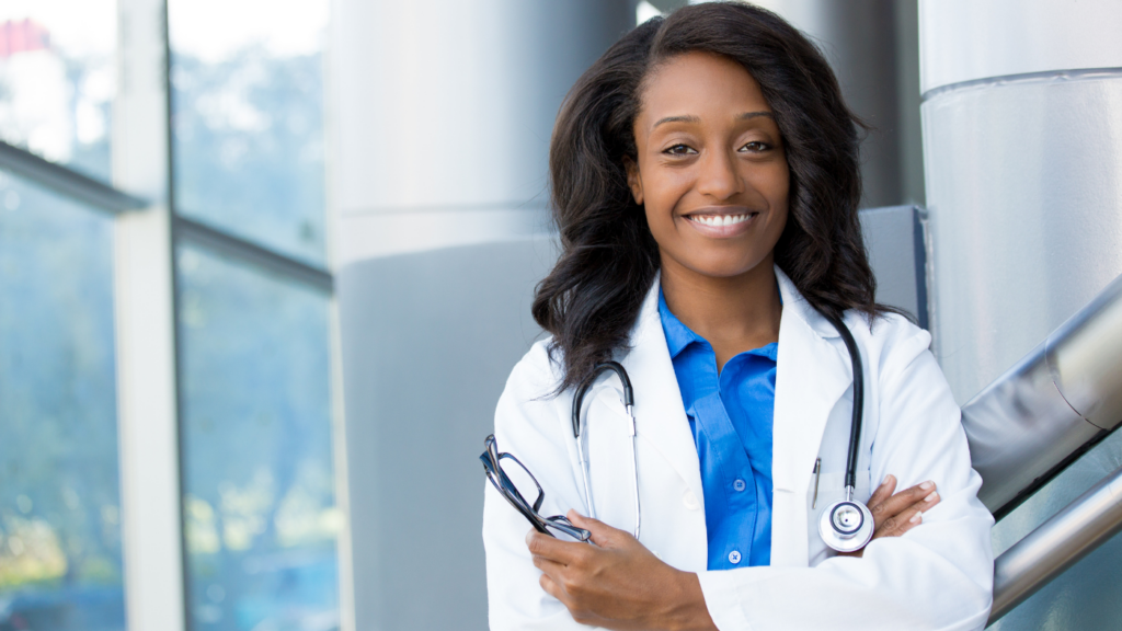 A smiling doctor in a white coat, holding eyeglasses and wearing a stethoscope, stands in a modern medical facility, exemplifying the difference between FNP and DNP expertise.