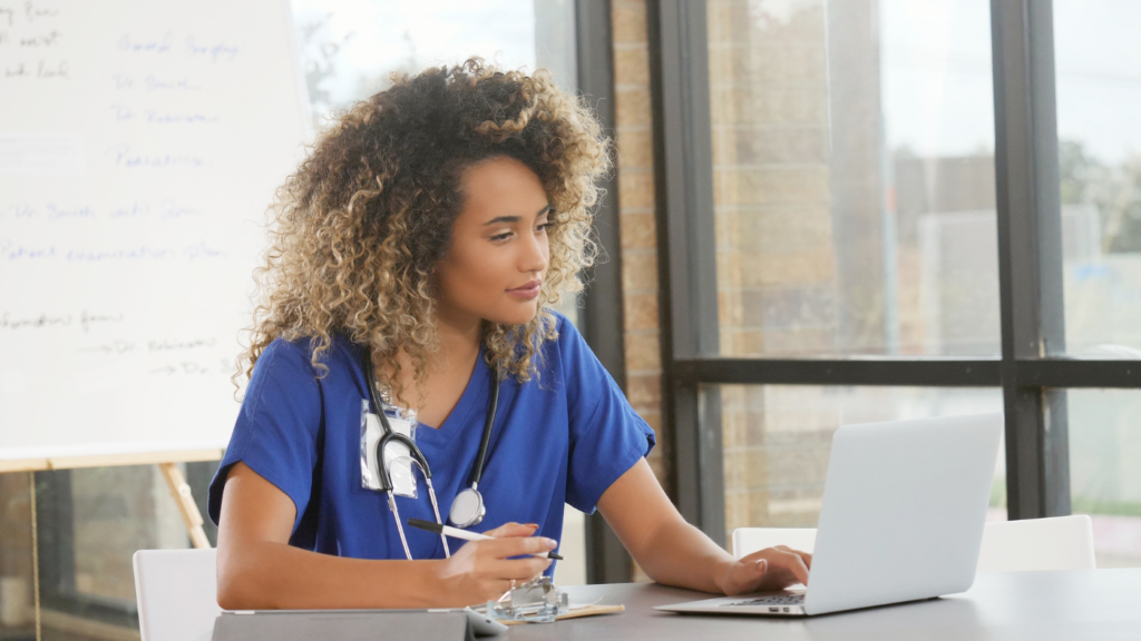 A person in medical scrubs, with a stethoscope around their neck, is seated at a table, looking at a laptop and writing on a clipboard, likely earning CE credits.