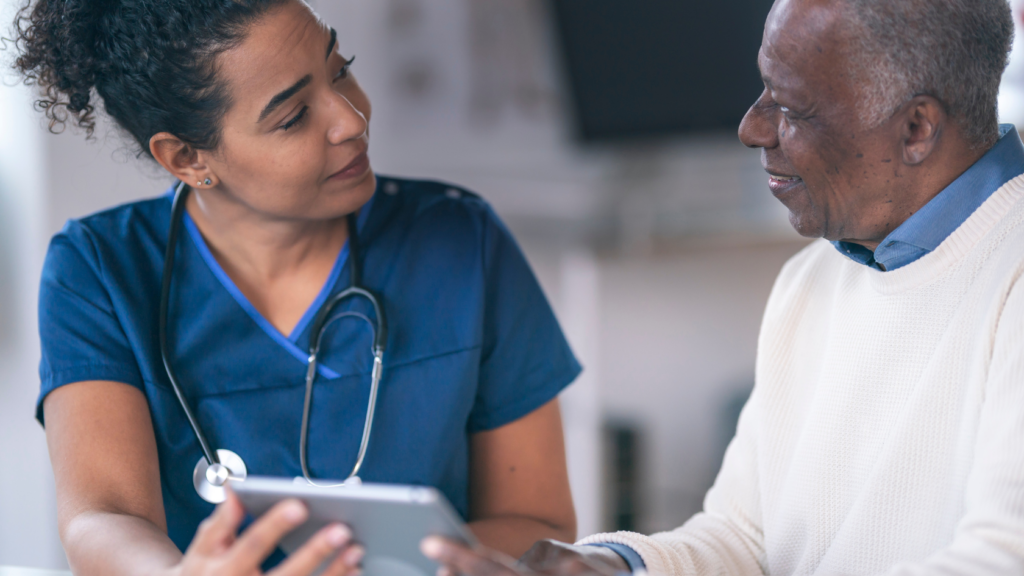 A nurse practitioner in blue scrubs shows a tablet to an older man in a white sweater. They appear to be engaged in a discussion, likely about health information, aiming for a positive impact on the patient's well-being.