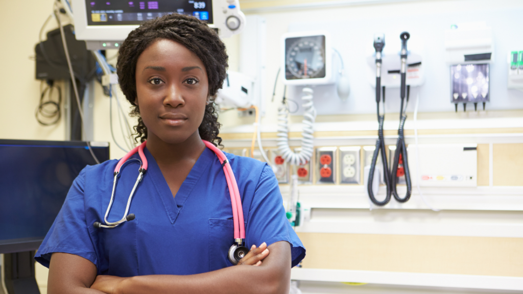 A healthcare professional wearing blue scrubs and a stethoscope stands with arms crossed in a medical facility, surrounded by medical equipment—everything you need to know to become an ENP-C.