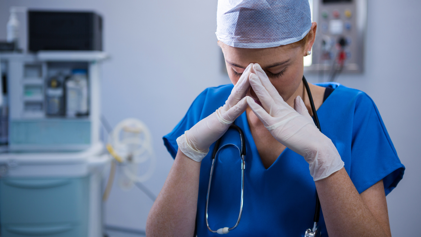 A nurse practitioner in blue scrubs and a surgical cap rests their head in their hands, appearing fatigued, highlighting the risk factors of burnout in a medical setting with equipment in the background.