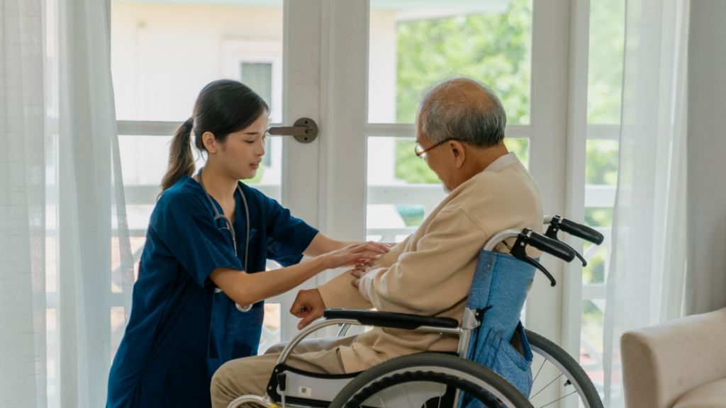 A young female Nurse Practitioner in blue scrubs tends to an elderly man seated in a wheelchair near a bright window.