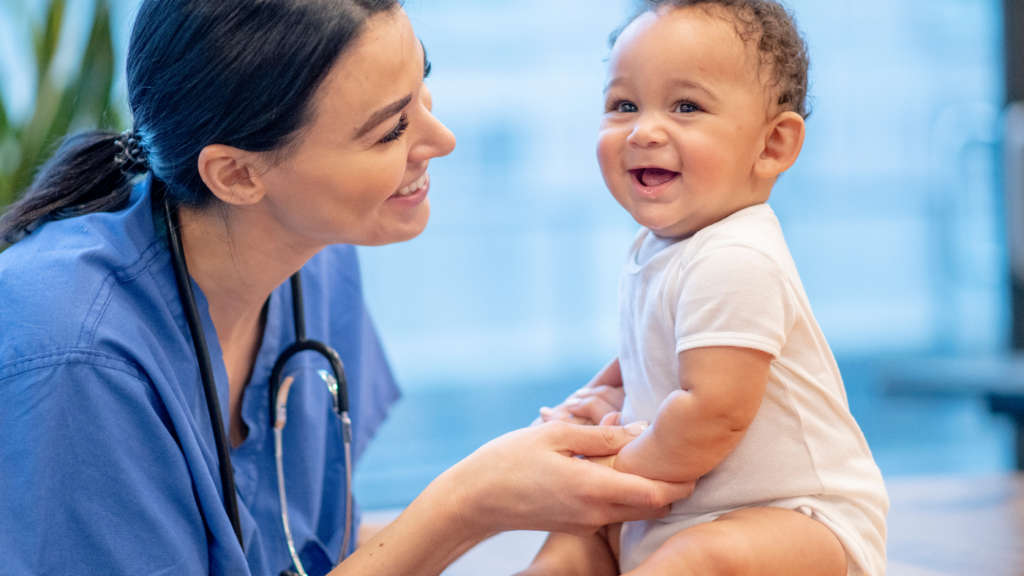 A Nurse Practitioner in blue scrubs smiles at a laughing baby sitting on an examination table. The baby, wearing a white onesie, giggles in delight.