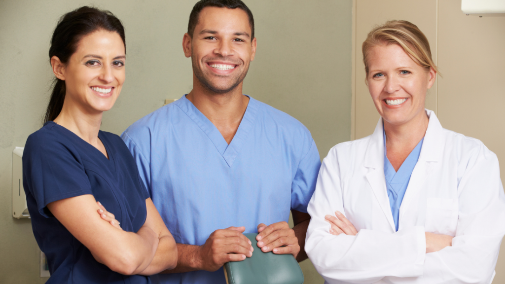 Three medical professionals, two women in dark blue scrubs and one man in light blue scrubs, stand smiling with arms crossed in a clinical setting, radiating confidence despite common struggles like imposter syndrome.