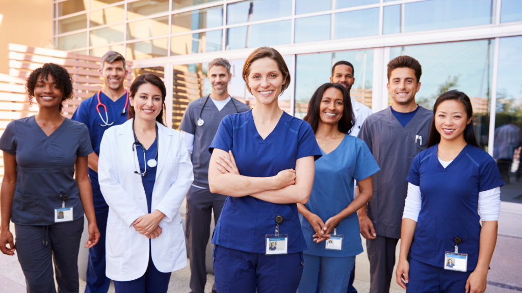 A group of smiling healthcare professionals, including doctors, nurse practitioners, and nurses, stand outside a medical building, exemplifying the broad scope of practice within their dedicated team.