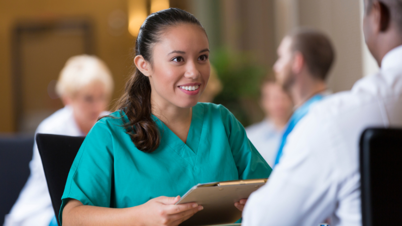 A woman in medical scrubs, likely a nurse practitioner, holds a clipboard and smiles while discussing expectations with another person in a healthcare setting. Other people are visible in the background.