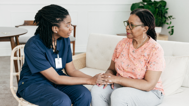 A nurse practitioner in blue scrubs sits holding hands with an older woman in a floral shirt, both seated on a couch in a well-lit room, embodying compassionate women's health care.