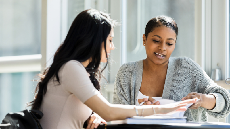 Two women sit at a table in a bright, modern setting, discussing papers and documents. One woman points at a document titled "NP Board Prep Do's and Don'ts," while the other looks on attentively.