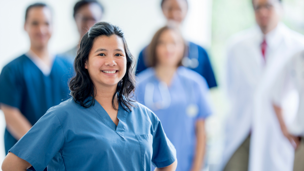 A woman in blue scrubs, embodying the confidence of a DNP graduate, smiles at the camera with a diverse group of healthcare professionals standing blurred in the background.