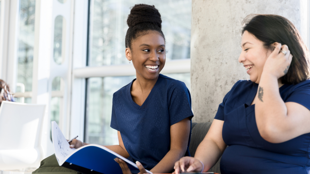 Two women in blue scrubs sit on a couch, smiling and holding documents about becoming a Nurse Practitioner.