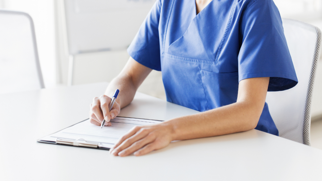 A person in blue medical scrubs is seated at a table, writing on a clipboard with a pen, focused on thriving in their NP job and avoiding burnout.