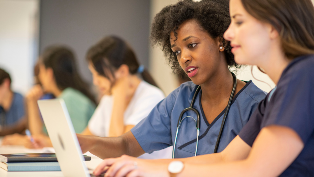 Two medical professionals in uniform, likely NPs, are using a laptop together in a classroom setting, with others working in the background. A stethoscope is draped around one professional's neck, highlighting their dedication to continuing education and earning CE credits.