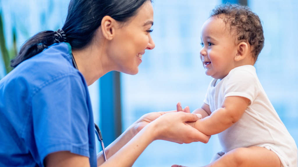 A nurse practitioner in blue scrubs interacts warmly with a smiling baby who is sitting up, holding the baby's hands during a primary care check-up.