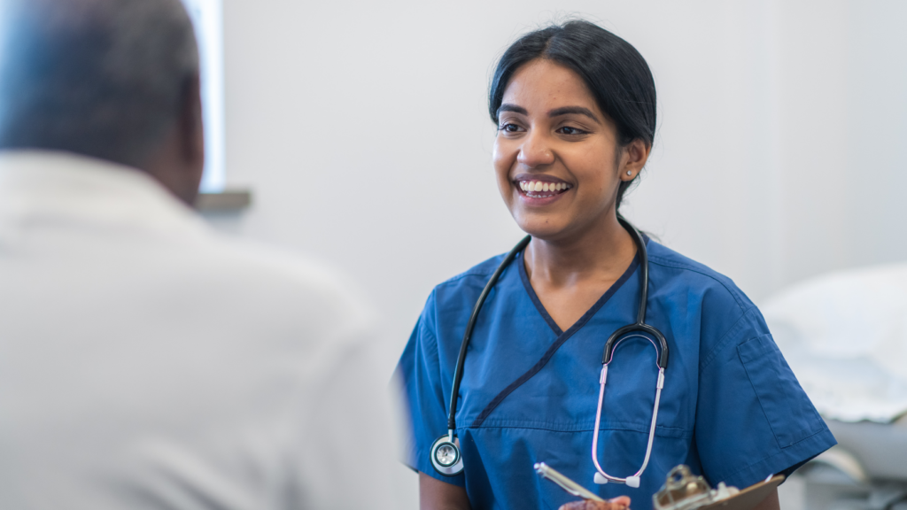 A nurse practitioner in blue scrubs, with a stethoscope around her neck, smiles while speaking to a seated patient in a medical setting, exemplifying the positive impact she has on her patients' lives.