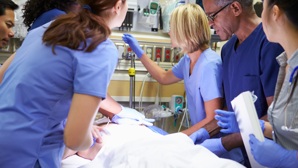 A medical team in scrubs surrounds a patient on a gurney, performing an emergency procedure. The lead nurse, an ENP-C, is calm and collected, directing the team with precision. In this critical moment, it's essential that everyone knows exactly what they need to know to ensure the best outcome.