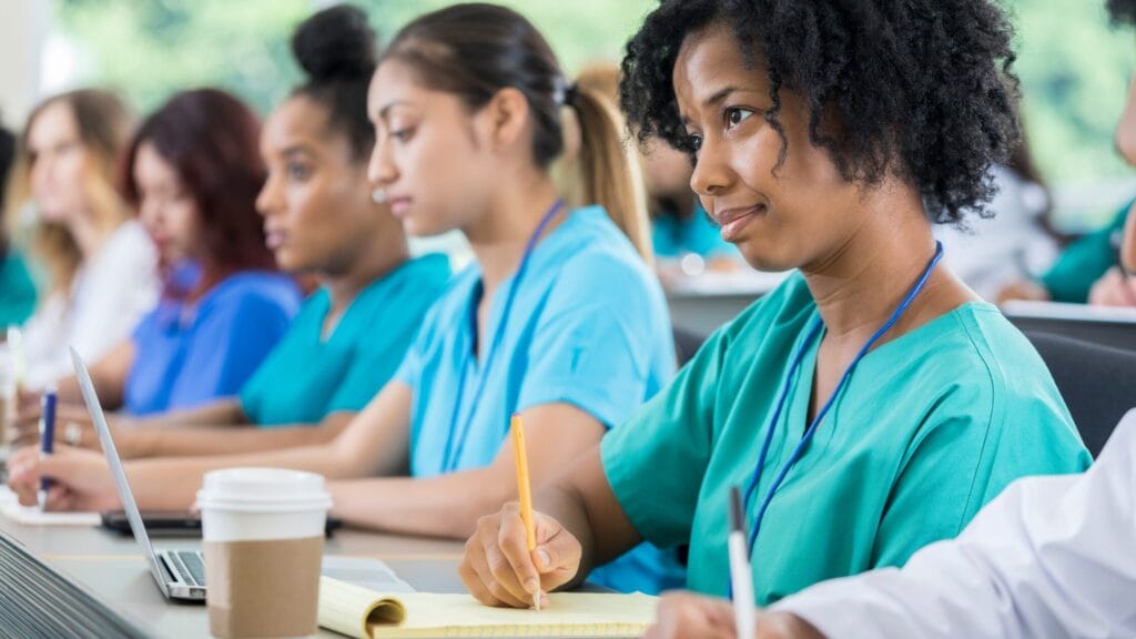 A group of nursing students in scrubs, aspiring to become confident nurse practitioners, sit attentively in a classroom, taking notes and using laptops.