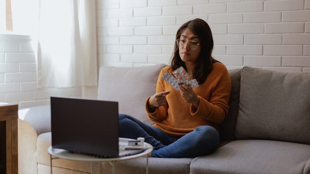 Telehealth is the future of modern medicine for nurse practitioners. In this Image, a woman sits on a couch with her legs crossed, holding blister packs of pills and pointing at them. A laptop is open in front of her on a small table, showcasing the ease of telehealth consultations in modern healthcare.