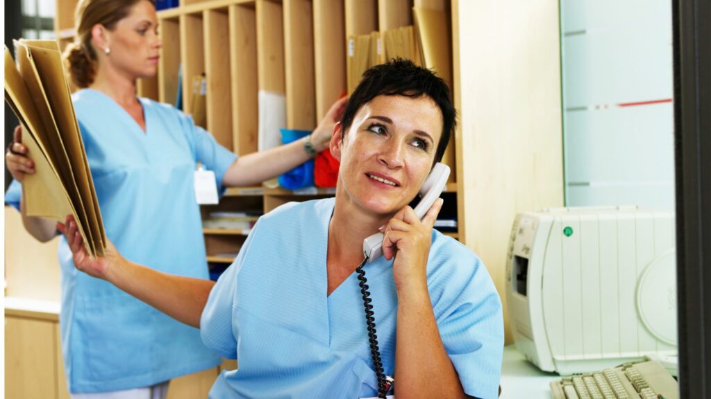 Two healthcare professionals in blue scrubs efficiently manage the reception desk. One is on the phone, handling files, while the other skillfully organizes paperwork. With stress-free strategies and patient load management, they maintain a seamless workflow amid computers and shelves in the background.
