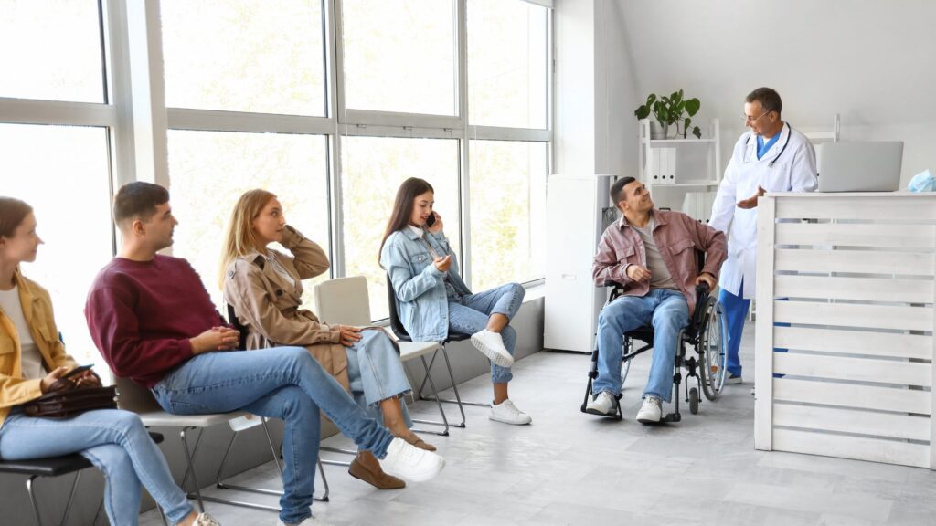 People sit in a waiting room; one person is in a wheelchair. A doctor, alongside new nurse practitioners, is speaking to them near the reception desk, showcasing their skills and dedication to patient care.