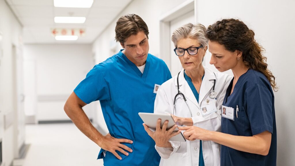 Three medical professionals, showcasing new skills, gather in the hospital corridor. Two nurse practitioners in blue scrubs join a colleague in a white coat as they discuss insights around a tablet.