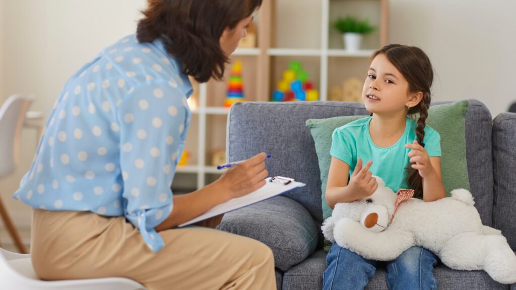 A girl sits on a gray couch, clutching a teddy bear, as she talks to a woman taking notes on a clipboard in what seems to be a thoughtful healthcare practice. Shelves filled with books and materials line the background, suggesting discussions perhaps tied to SDOH.