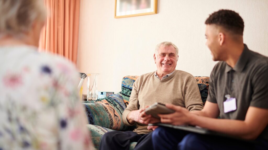 An elderly man and a younger man sit on a couch, smiling and discussing healthcare. The younger man holds a tablet, perhaps reviewing SDOH insights. Another person is partially visible in the foreground.
