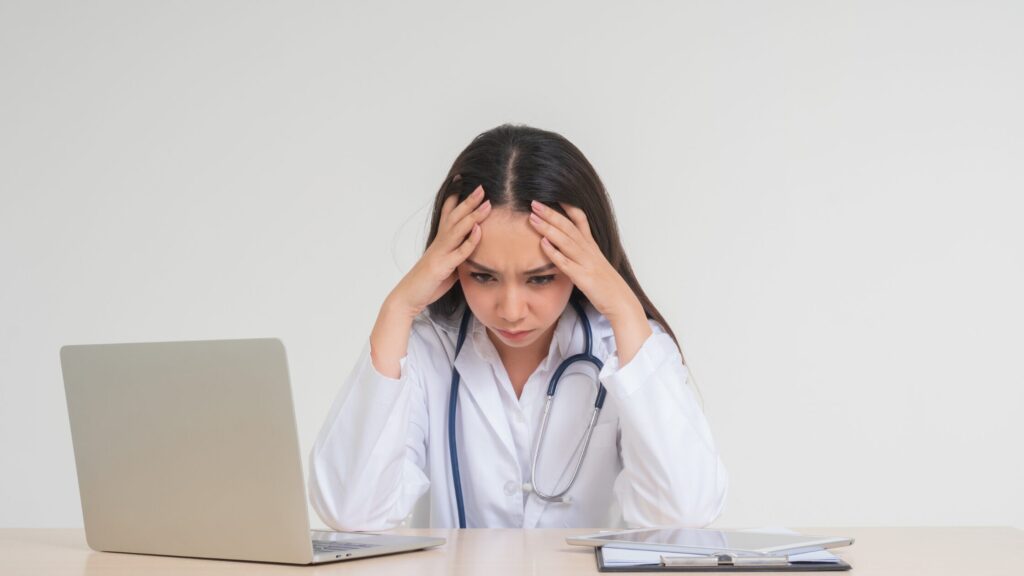 A Nurse Practitioner in a white coat sits at a desk, looking stressed with hands on their head, surrounded by a laptop and insurance and billing documents.