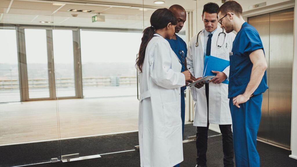 Four medical professionals, including a nurse practitioner, stand together in a hospital hallway, discussing documents while demonstrating the importance of interdisciplinary teams and collaborative care.