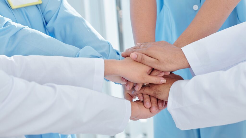 A group of healthcare professionals, including a nurse practitioner, stacking their hands together in a gesture of teamwork and unity, highlighting the strength of interdisciplinary teams.