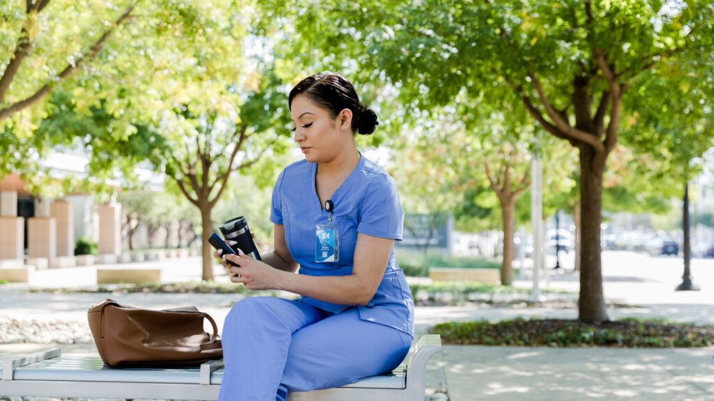 A woman in blue scrubs sits on a bench outdoors, practicing time management as she looks at her phone with a brown bag beside her, surrounded by green trees and sunlight.