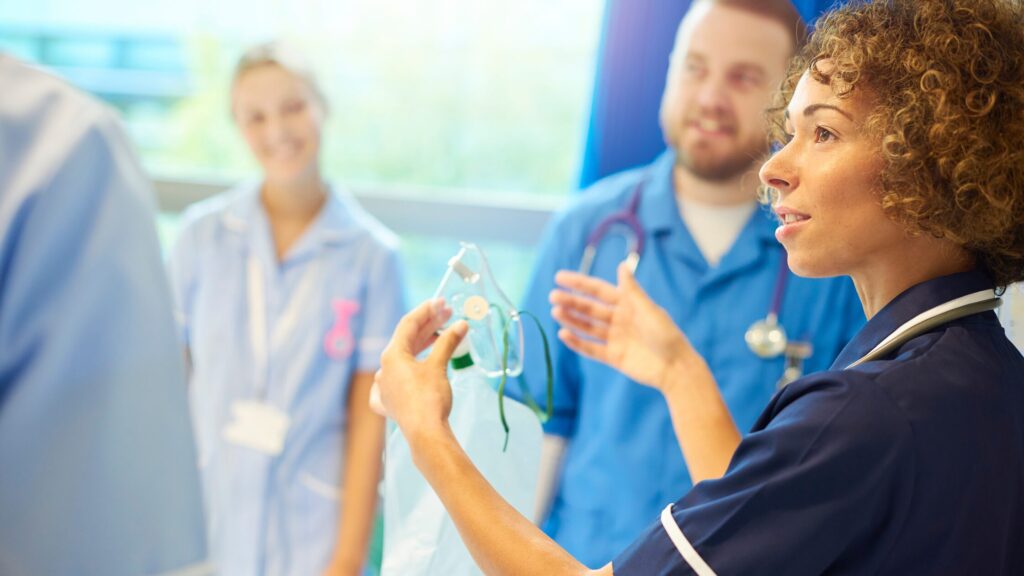A nurse with strong leadership skills holds up an oxygen mask and speaks to a group of medical professionals in scrubs, who are listening attentively in a hospital setting.