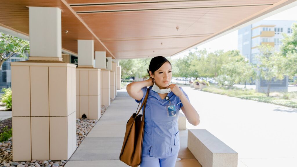 A woman in medical scrubs walks outside a building, carrying a tote bag and adjusting her face mask around her neck, reflecting the time management skills needed for busy healthcare professionals.