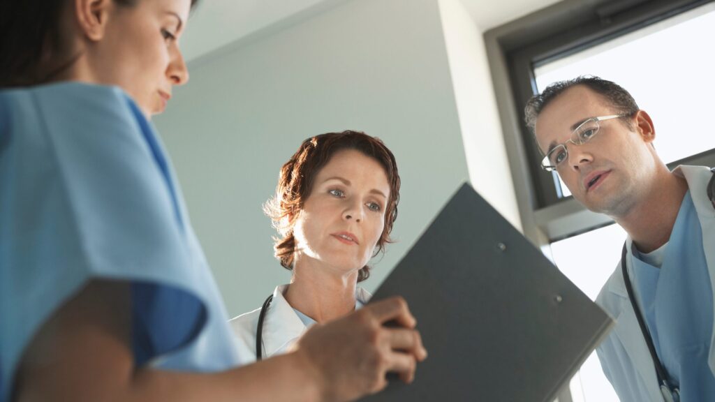 Three medical professionals, including nurse practitioners in scrubs and lab coats, review information on a clipboard in a brightly lit room, highlighting leadership skills and teamwork essential in primary care settings.