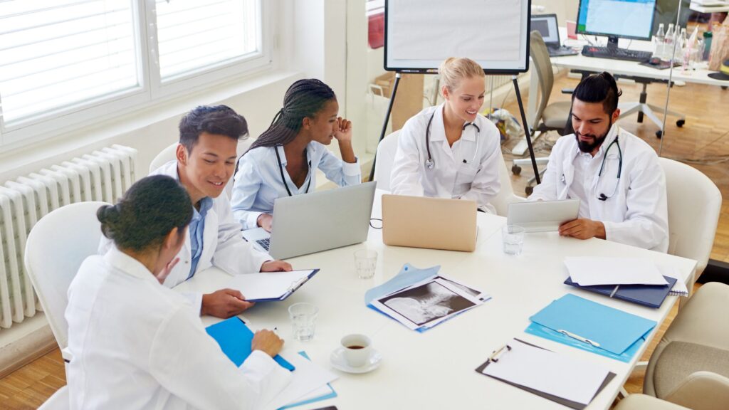 Five providers in lab coats sit around a table with laptops, documents, and medical images, engaged in a group discussion that highlights teamwork in a well-lit office.