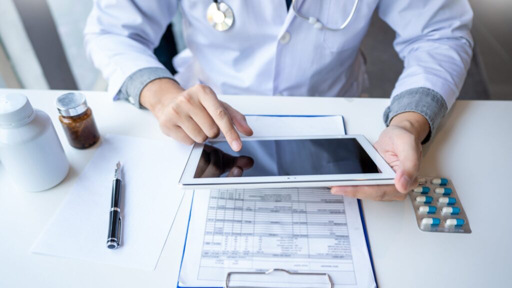A doctor wearing a white coat uses a tablet at a desk with medical forms, pills, a pen, and a medicine bottle—an ideal scene to discuss NP mistakes and share nurse practitioner tips for new NPs in clinical practice.